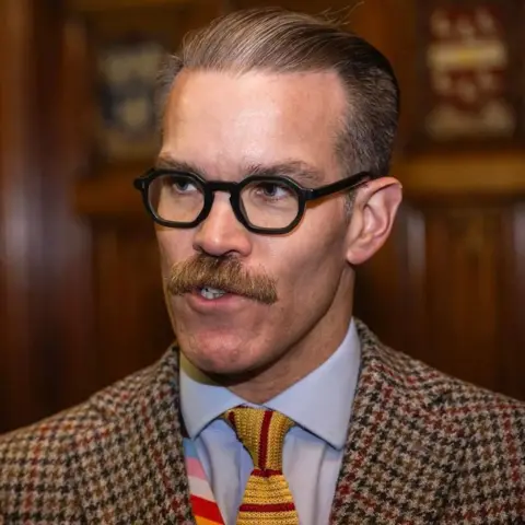 House of Commons A man with graying, swept back hair and brown mustache is wearing a checked jacked with a yellow and red woven tie and pale blue shirt. He has thick, black octagonal-framed glasses. 