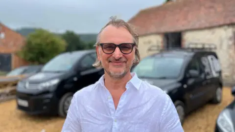 George Carden/BBC Adrian Atwood wearing a white shirt standing in the yard of the education centre which is a set of converted barns