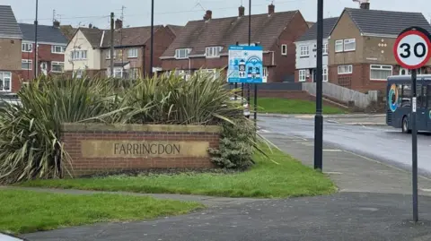 A brick sign on a street saying Farringdon. It is on a patch of grass and there are plants behind it. There are houses in the background.