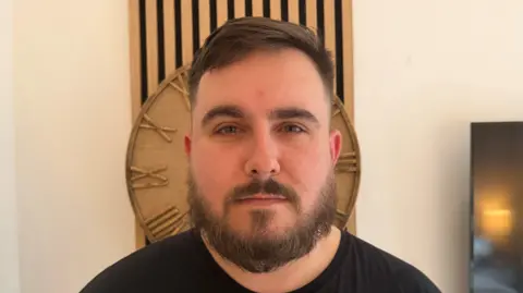 Chris Grundy Close up image of a young man, head and shoulders, with full beard and moustache, wearing a black t-shirt. He has a serious look on his face and there is a wooden clock directly behind his head.