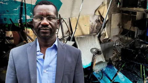 Mohammed Idris has black hair and is wearing glasses, a blue blazer and light blue shirt. In the background is the interior of his cafe after it was burnt. 