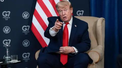Donald Trump sat in a cream coloured armchair points with his finger. Behind him is a G7 logo and Unites States flag.