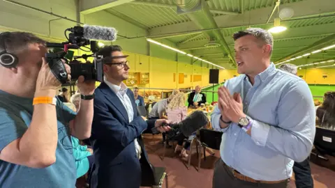 Man wearing pale blue shirt being interviewed by a suited journalist, holding a microphone, and a cameraman wearing a blue t-shirt, in a sports hall with people sat down in the background