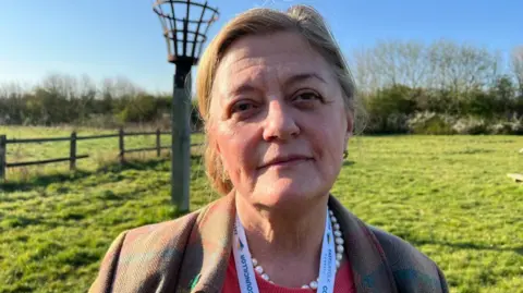 Julia Ewart is standing outdoors in a grassy field on a sunny day. She is wearing a plaid-patterned coat in muted tones over a coral-coloured top, accessorized with a pearl necklace and a East Suffolk Council lanyard. Behind her there is a wooden fence and a tall metal basket-like structure on a pole. 