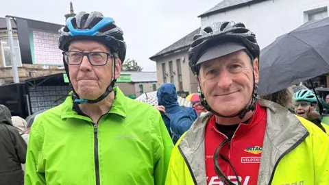 Two men wearing cycling helmets stand side by side outdoors on a rainy day. One wears a bright green waterproof jacket, while the other wears a red cycling jersey under a light rain jacket. Several umbrellas, cyclists, and buildings are visible in the background