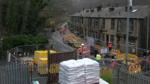 A picture showing a road closed off by road works. Next to the roadworks are a row of houses and on the other side of the road is a business premise.