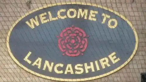 Oval sign saying "welcome to Lancashire" with a red rose in the middle. the writing is gold on a black background