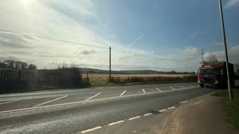 A lorry driving along a road in the foreground alongside a field behind a fence in the background