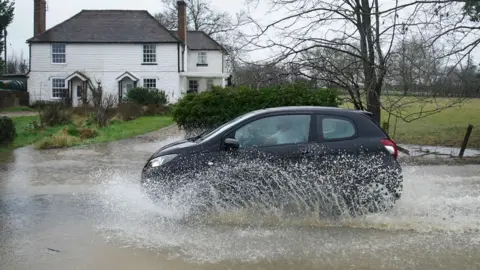 PA Media A car passes through a flooded road with a white house in the background.