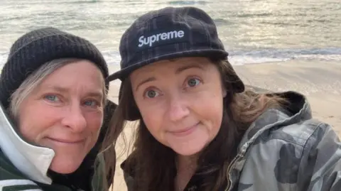 Una Mullally Two women looking towards the camera, one is wearing a black woolly hat the other is wearing a black cap that says SUPREME in white letters. They are standing on a beach.