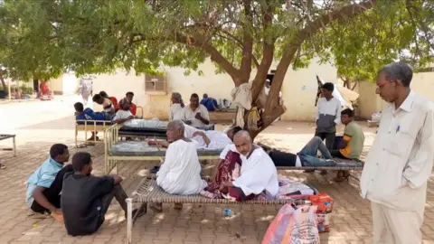 Groups of patients sit on beds and on the floor outside the hospital.