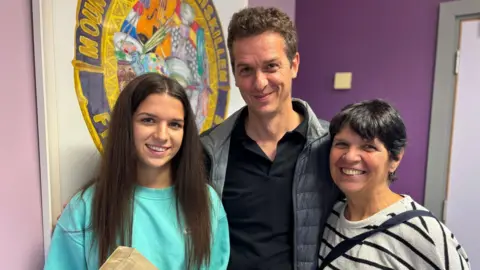 A young woman with long dark brown hair, with her parents