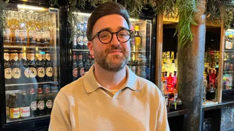 A man with dark hair and glasses smiles while standing behind a bar, with bridges full of beer behind him 