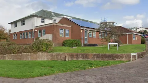 A street-view of Honiton Surgery in Devon. It is a red-brick building with a small wall surrounding it. There is grass and bushes in front of the building and two white street signs on the right. 