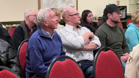 A group of people are sitting down and looking at Steve. One man is asking a question. He is wearing a purple zip up jumper and has short grey hair.