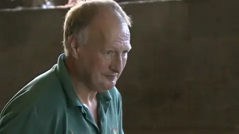A screenshot from a TV interview with David Knipe. He has short receedeing white hair and blue eyes. He wears a green polo shirt and is inside one of his poultry sheds. He is looking at something out of shot to the side.