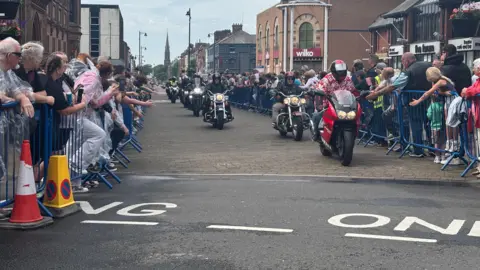 Hundreds of people line up behind barriers in Barrow as Dave Day 2 goes ahead with motorcyclists in the road who have travelled from London. The biker in front is wearing a red Hawaiian-style shirt.
