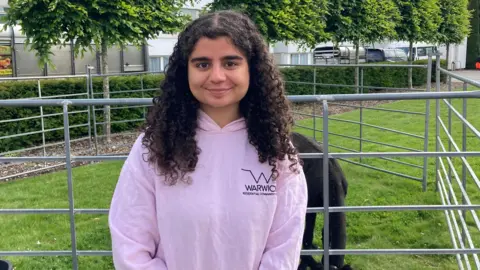A young woman in a pink Warwick University hoodie and long curly black hair is standing in front of a pen where a black Shetland pony is eating the grass beneath. There is a row of trees in the background.