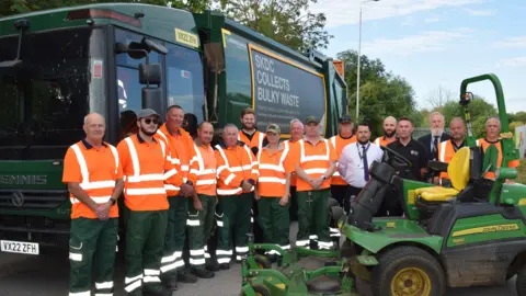 A group of uniformed workers in hi-vis orange tops, standing in front of a green bin lorry. There is a lawnmower in the foreground. There are trees in the background. The sky is blue with light clouds.
