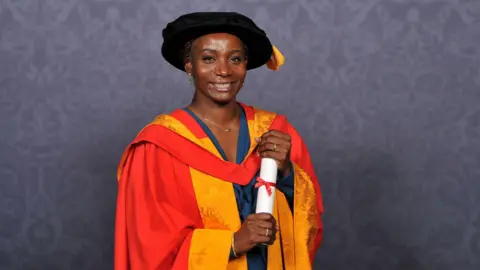 University of Wolverhampton Fatou Baldeh MBE is pictured holding a white scroll and smiling. She is dressed in a long orange and red graduate cloak and has a black hat.
