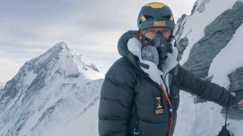 Tim Howell is standing on a snowy mountain. A mountain ridge is to the left of the image. He is wearing outdoor gear and an oxygen mask on his face. He is to the right of the image, staring into the camera.