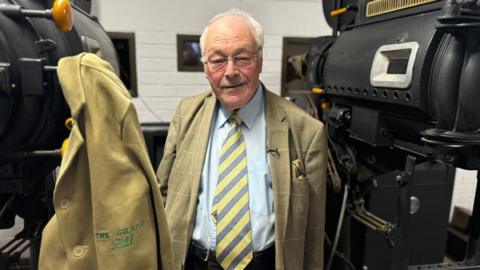 Bill Mather, dressed in a beige jacket with a yellow tie, stands next to projectors with his old projectionist jacket hanging up next to him.