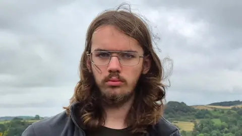Handout Tobias has long hair and glasses and is standing outdoors in front of rolling countryside under a cloudy, overcast sky.