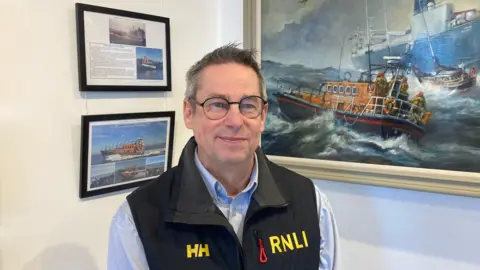 David Gillingwater wearing a black RNLI jacket, with yellow branding and lettering, stood in front of pictures of lifeboats mounted on the wall.