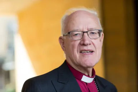 Getty Images Portrait of George Carey, who has balding white hair and black-framed glasses. He is wearing a purple clerical shirt with white clerical collar and a black pinstripe jacket. He is pictured outside in front of a yellow-painted wall. 