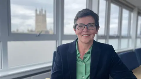 NHS Gloucestershire Mary Hutton, wearing a green blouse and navy blazer. She has cropped hair and is wearing glasses, while smiling at the photographer.
There are windows behind her and it looks like Gloucestershire Cathedral in the background, which is out of focus.