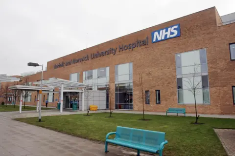 An entrance to a hospital with brown bricks and blue benches in the foreground, with grass and trees around them.
