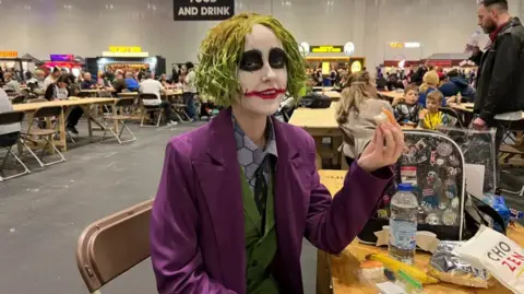 Thomas Copeland / BBC News A young woman sits at a table eating sushi, dressed as the Joker with scruffy green hair and clown makeup, wearing a purple suit with a green vest.