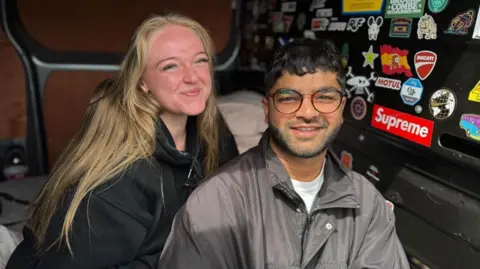 Adam Deen and his partner Shania Evans sit in a van with lots of stickers on the wall. Shania has long blonde hair and wears a black hooded jumper. Adam has short dark hair, he wears glasses and a grey jacket.