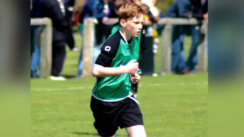 Anna Kilpatrick Isaac playing football as a 12 year old boy. It is an action shot of him running on a sunny day. Blurred in the background are the supporters watching the game. 