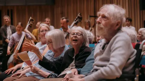 Plaster Care home residents sit in the audience as the watch an orchestral concert at the Bristol Beacon. There are musicians behind them playing cellos.