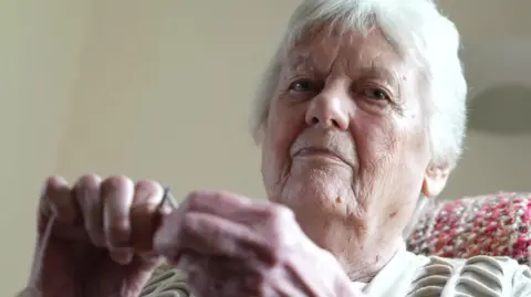 Granny H, with white hair, sits in a chair as she knits