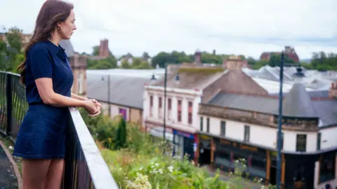 STV Studios Factual/BBC A woman wearing a navy blue, short-sleeved shirt and denim shorts, standing by a railing overlooking a town with rooftops, shops, and greenery in the background