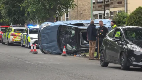 Supplied An overturned car - part on a road, part on the pavement - between parked cars and in front of two police cars. Police officers are talking to a man next to the car.