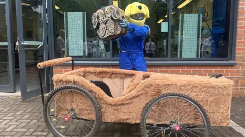 BBC/ELLIE COLTON A soapbox kart covered in brown fur-like material with bicycle wheels. A woman, Ellie Colton, wearing a crash helmet and blue boiler suit, holds up a part of the kart bearing the face of a pug.