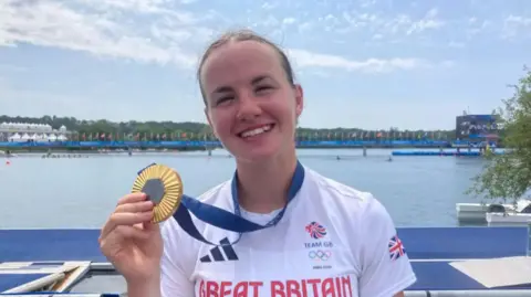 Lauren Henry showing her Olympic gold medal in Paris. She is wearing a white Team GB shirt.