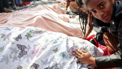 REUTERS/Ricardo Moraes A woman touches a body, covered with a cloth, with her hand. She looks into the camera, she looks distraught. Another woman is leaning on the first woman's shoulder. Many other bodies can be seen behind the body the woman is touching. They are all covered with cloths and arranged next to each other