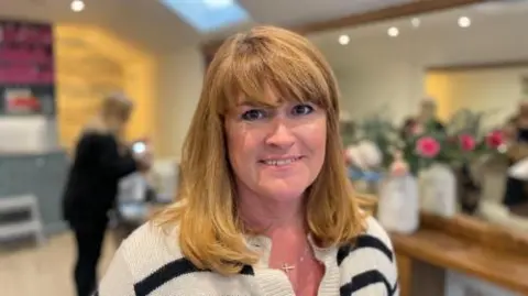Jayne Prigent stands in the centre of one of her hair salons. She was a striped cardigan and has long blonde hair with a fringe. There's a client and a member of staff in the background, and a vase of flowers on one side