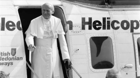 Getty Images A black-and-white image of a man in white ecclesiastical robes with crucifix round his neck, holding a metal rail as he emerges from a helicopter, one hand poised to wave. He is bald and wearing a white skullcap or zucchetto.