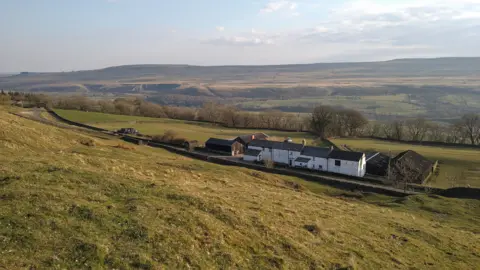 Emma Spry A long white farmhouse in four sections is set at the bottom of a steep hill alongside a single track rural road. Behind it are hills of moorland 