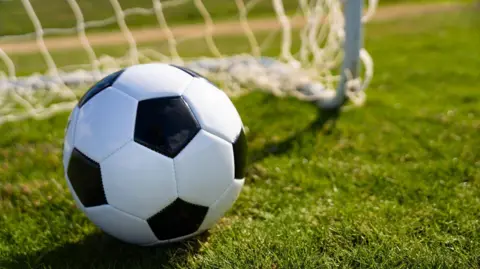 A stock image of a black and white patterned football, on green grass with a white net and goalpost visible in the background