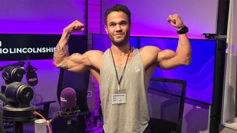 Donald Sweeting standing with his arms aloft in the BBC Radio Lincolnshire studio. He has short dark hair and beard, tattoos on his right arm and is wearing a grey vest