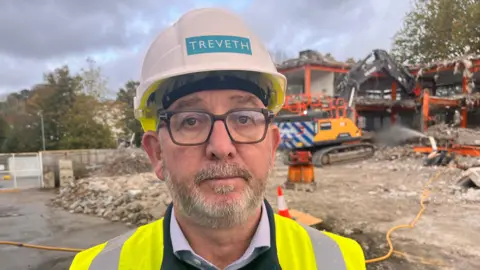 A man with a short beard and glasses stands at the demolition site with bulldozers behind him. He wears a hard hat and hi-viz jacket.