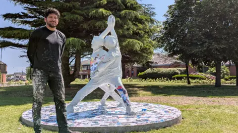 Helix Arts Aaron Li-Hill standing in front of the sculpture. He has dark, short, curly hair and is wearing black clothes which are covered in white paint.