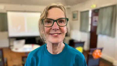 A women with short blonde hair, green rimmed glasses and a blue t-shirt. She is looking just above the camera and smiling. 