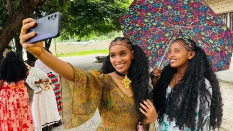 Girmay Gebru/BBC Two girls wearing colourful patterned dresses with a colourful umbrella smile and pose whilst taking a selfie. Their hair is in braids. They are outside and there are some women in dresses in the background as well as trees - Mekelle, Ethiopia. Sunday 24 August 2025.